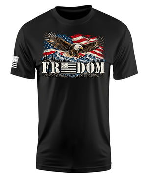 A black short-sleeve crewneck t-shirt featuring a patriotic graphic on the chest of an eagle flying over a mountain range beneath a waving American flag, with the word 'FREEDOM' printed below in white block letters. A small black-and-white American flag is printed on the right sleeve.