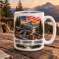 A white ceramic mug with a detailed patriotic mountain landscape print, featuring an American flag, rifle, coffee pot, and pine trees, with the text 'COFFEE + COUNTRY + CARBINES'. The mug is placed on a rustic wooden table with a blurred mountain sunset background.