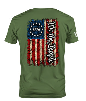 An olive green short-sleeve t-shirt shown from the back, featuring a distressed American flag graphic with the text 'We the People' in gothic font and '1776' inside a circle of stars.