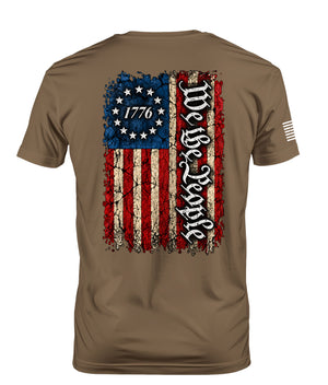 A brown short-sleeve t-shirt shown from the back, featuring a distressed American flag graphic with 'We the People' written vertically in black gothic lettering and '1776' inside the stars.
