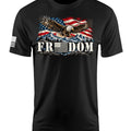 A black short-sleeve crewneck t-shirt featuring a patriotic graphic on the chest of an eagle flying over a mountain range beneath a waving American flag, with the word 'FREEDOM' printed below in white block letters. A small black-and-white American flag is printed on the right sleeve.
