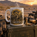 A white ceramic mug with a yellow and black Gadsden flag snake graphic sits on an olive green military-style ammo crate. The scene is set outdoors in a desert landscape at sunset, with a rifle, sunglasses, and tactical gloves nearby.